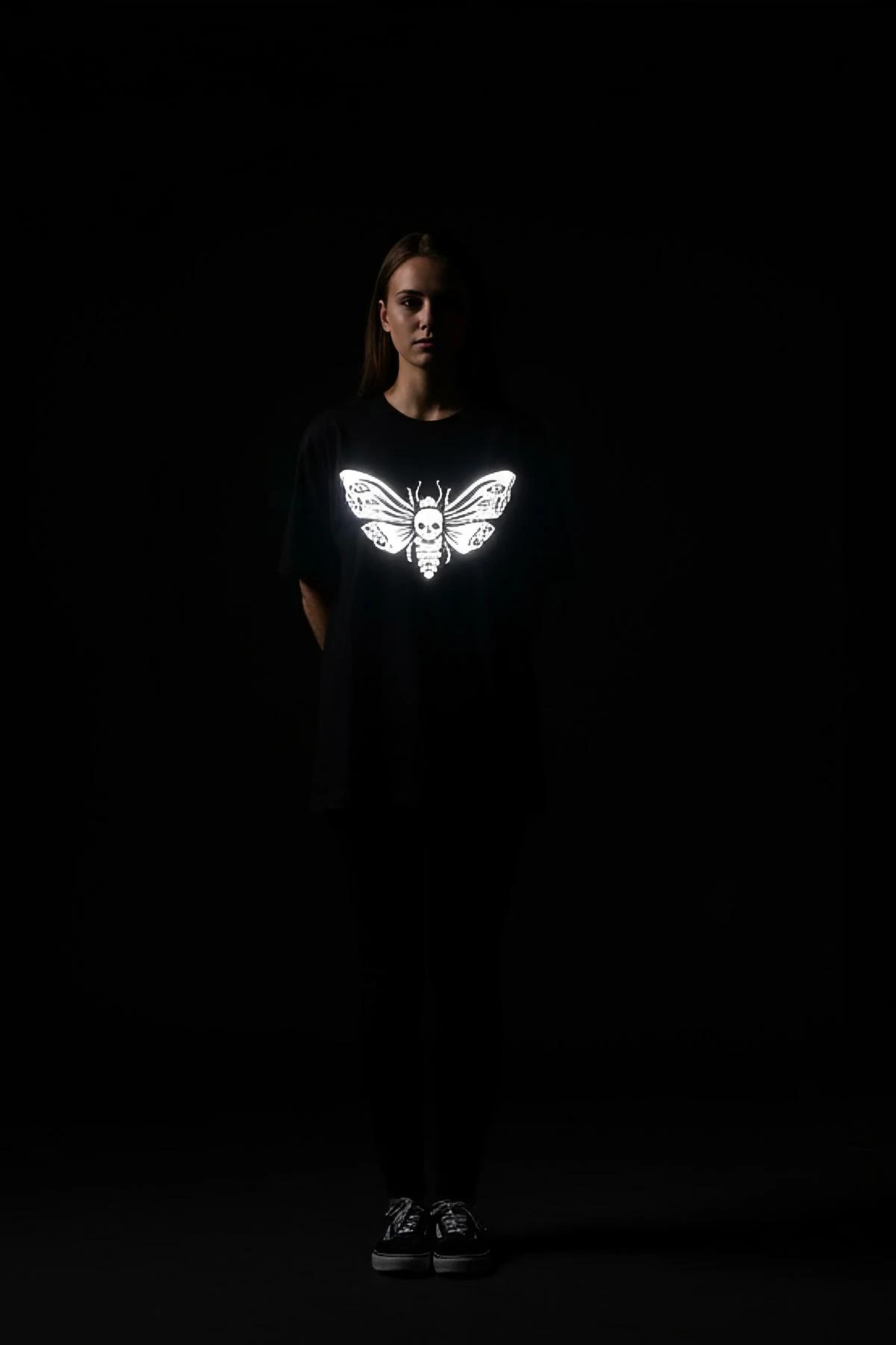 Reflective death's head moth black oversize t-shirt glowing bright white under flash, worn by a woman in a dark room