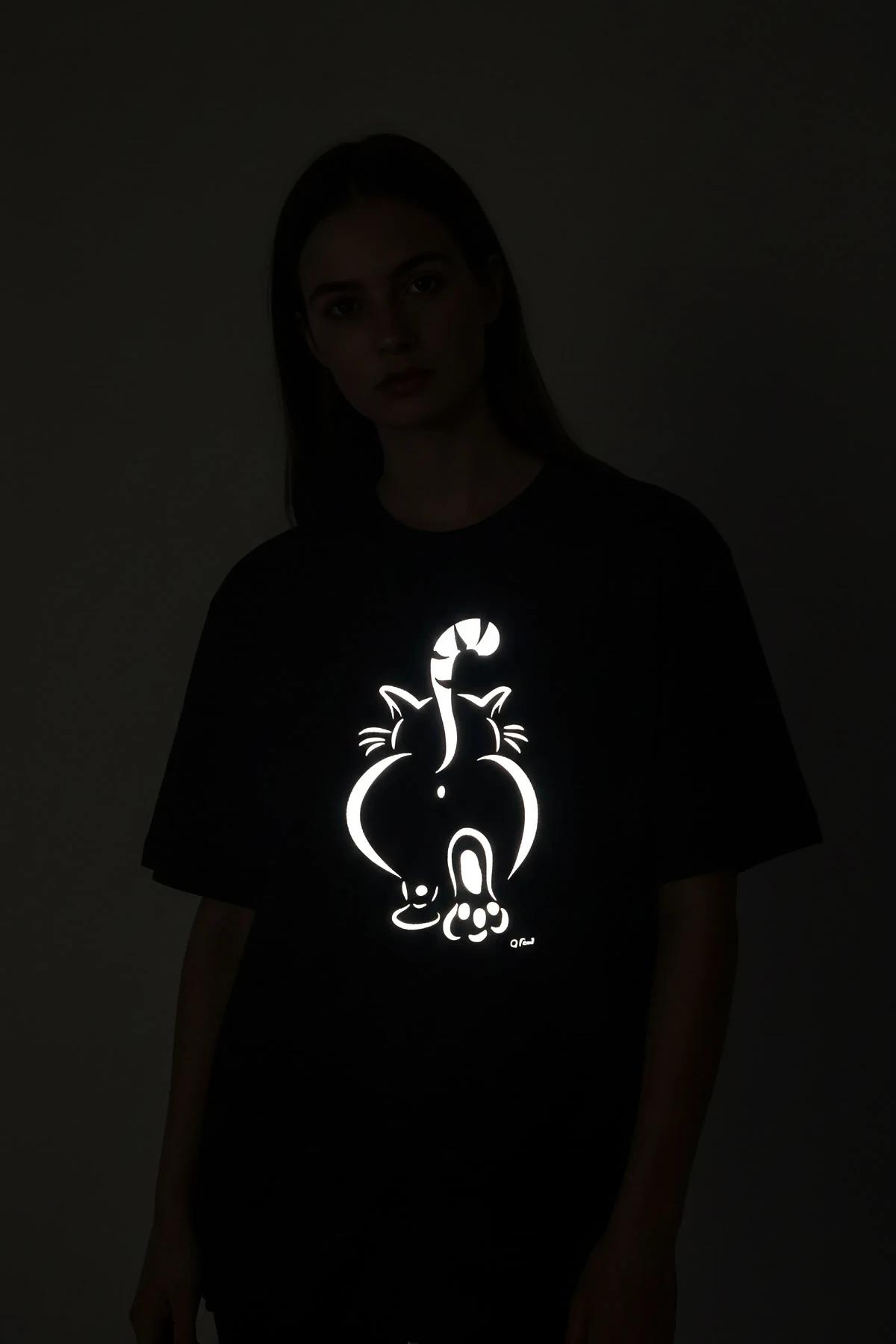 Reflective cat figure oversize black t-shirt glowing white cartoon cat-from-behind print under camera flash