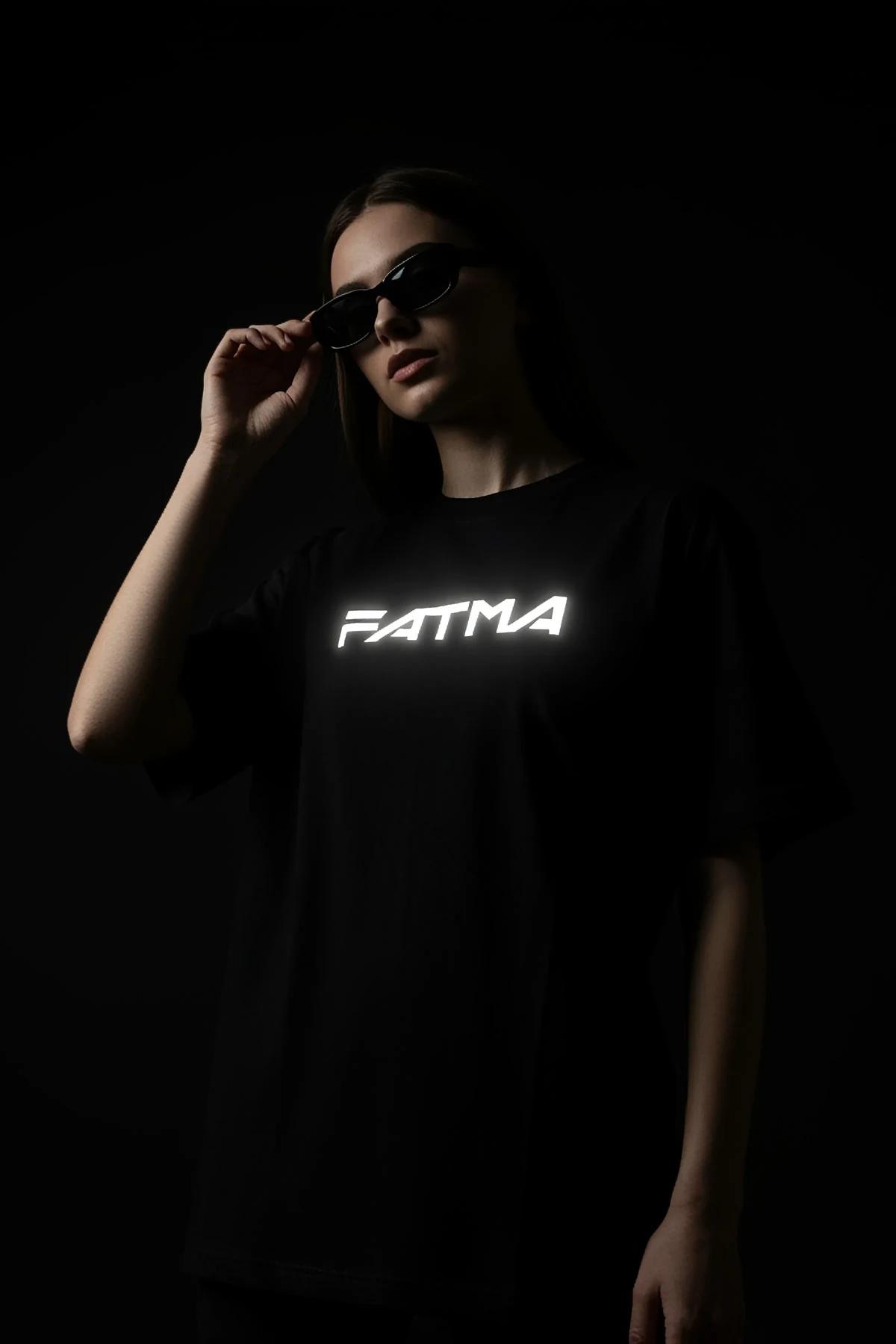 Black oversize t-shirt with reflective FATMA name print glowing white under camera flash, worn by woman in dark studio