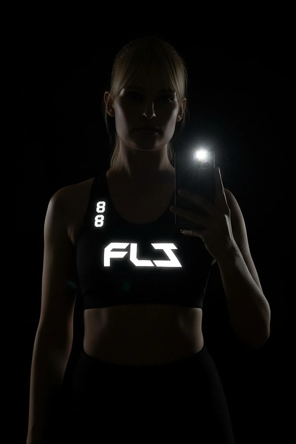 Black racerback sports bra with white retroreflective FLJ lettering glowing on chest under camera flash