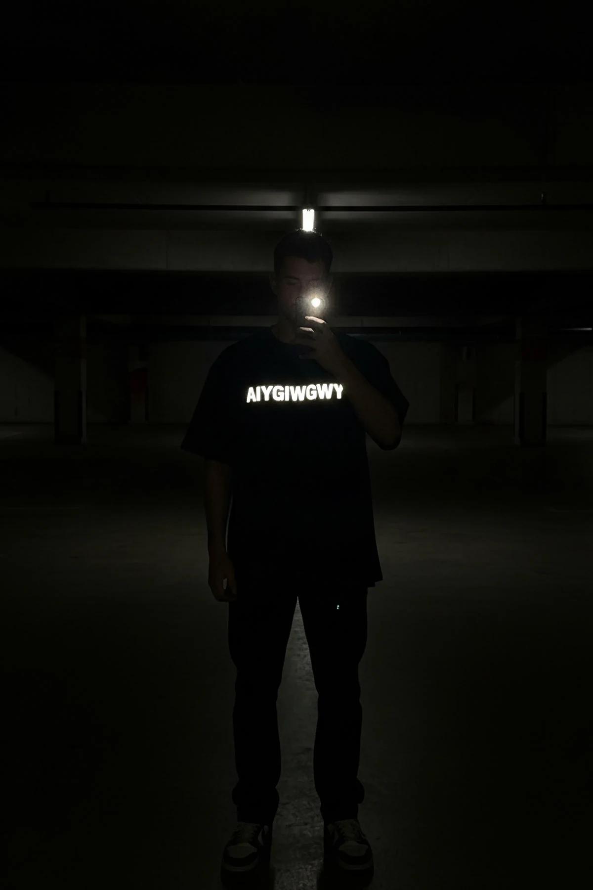 black reflective oversize t-shirt with AIYGIWGWY slogan glowing white under flashlight in dark parking garage