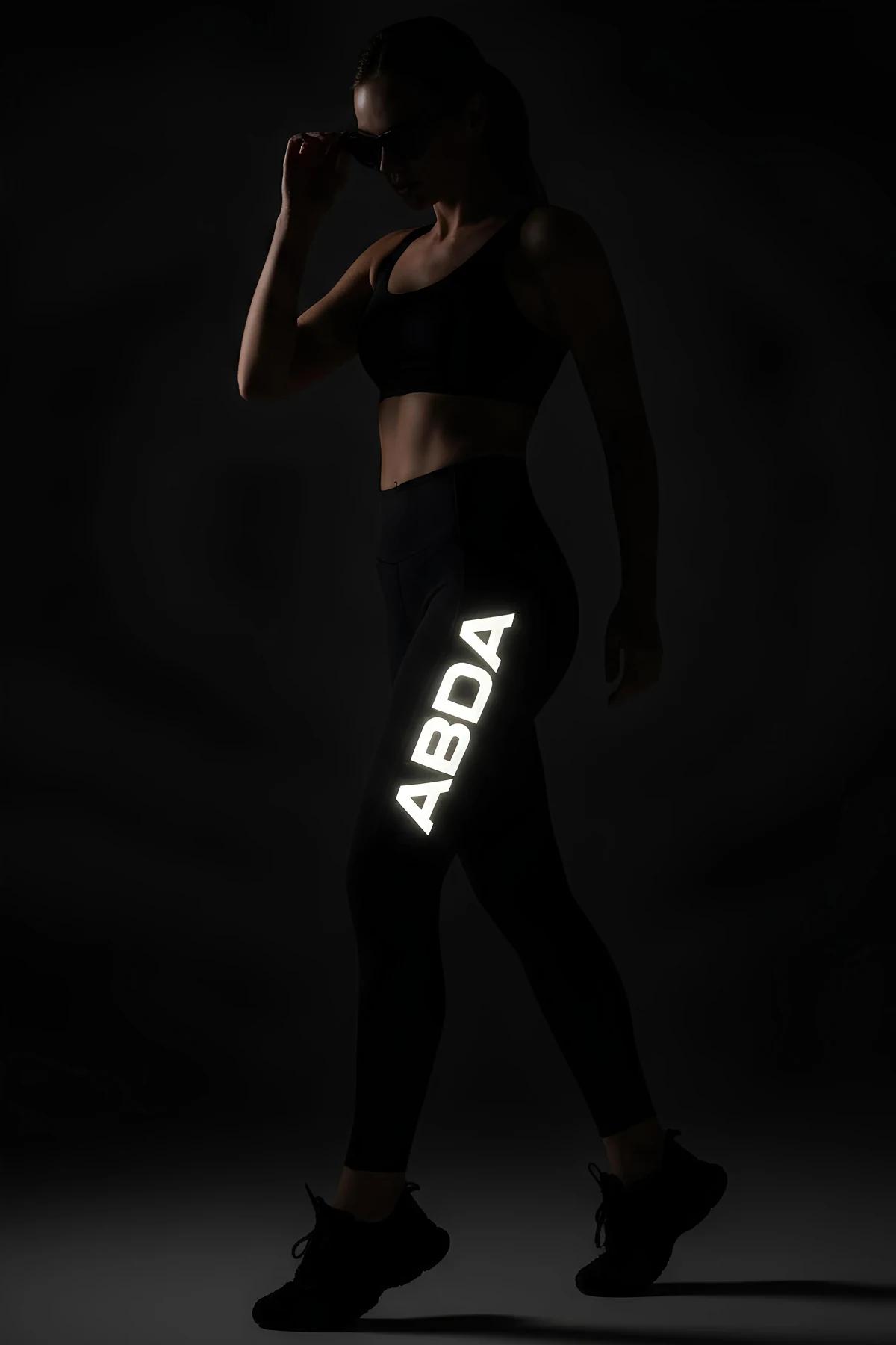 Black high-waist leggings with large reflective ABDA letter print glowing white on outer right thigh in dark studio