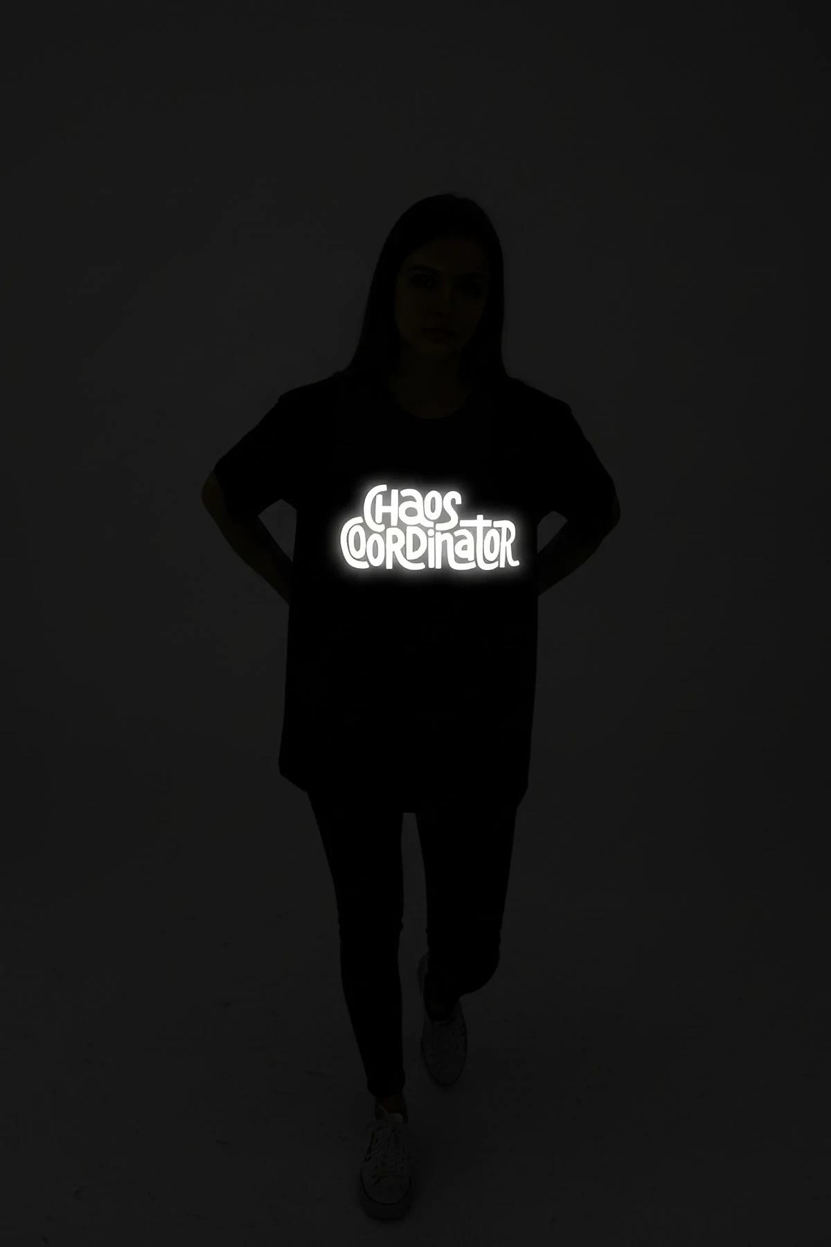 Black oversize t-shirt with bright white reflective CHAOS COORDINATOR front print glowing under direct light in dark setting