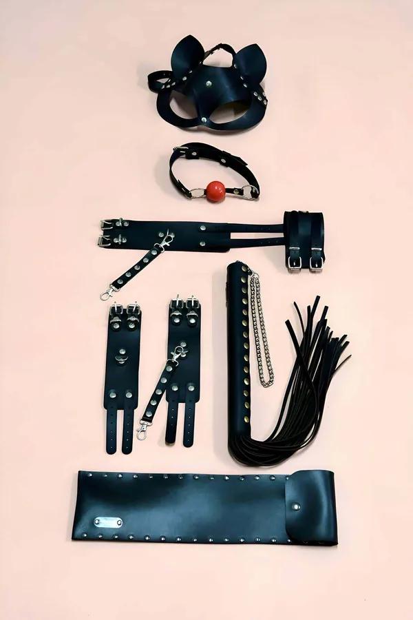 Black faux leather costume accessory set with cat mask, ball gag, spreader bar cuffs, flogger, and wide studded cuff on pink 