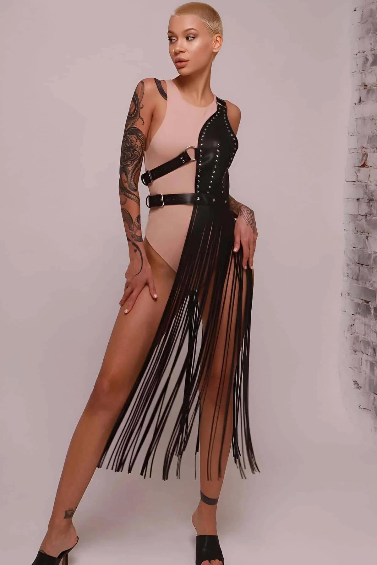 Black faux leather asymmetrical fringe harness skirt with silver stud trim and side buckle, worn over a nude bodysuit