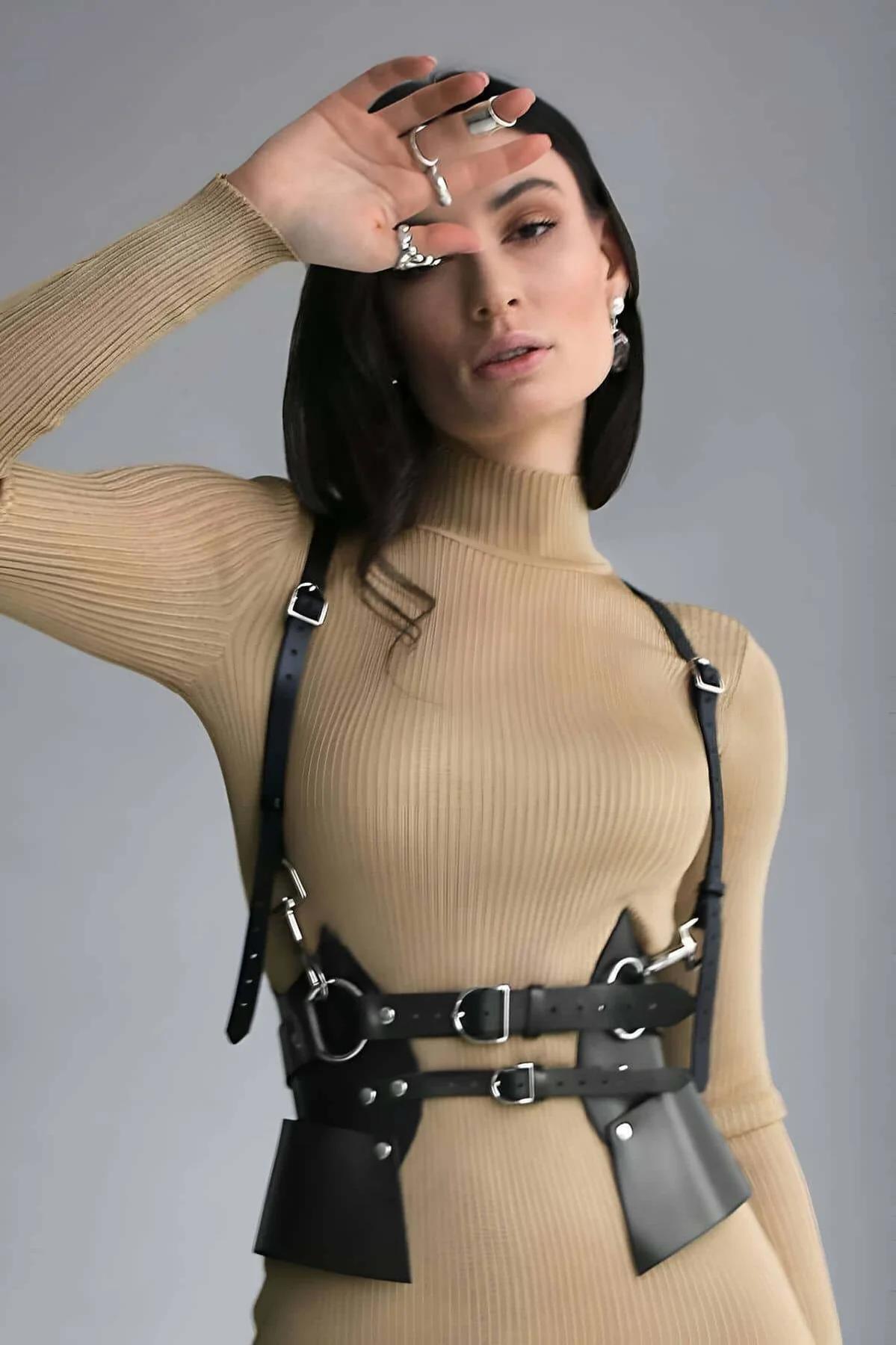 Black faux leather sculpted body harness with wide waist panels and silver buckles worn over camel ribbed turtleneck