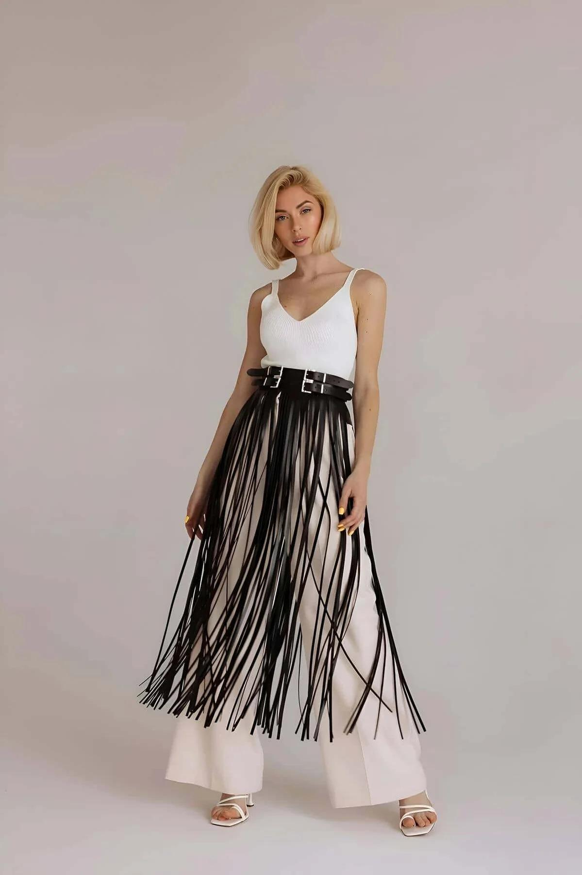 Black faux leather fringe belt skirt overlay with dual silver buckles worn over white wide-leg trousers, long fringe strands