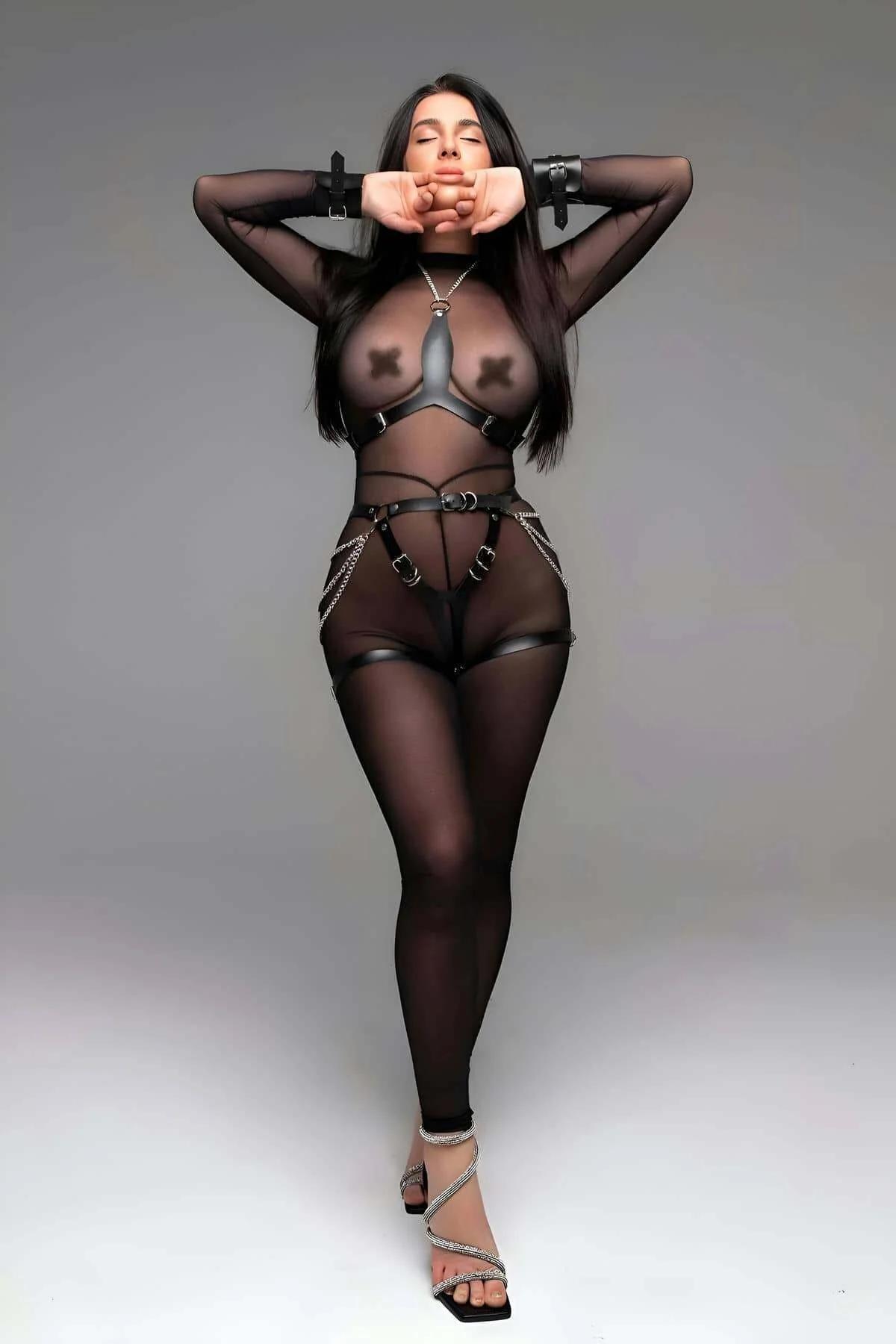 Black faux leather full body harness set with chest O-ring straps, silver hip chains, and wrist cuffs styled over sheer mesh