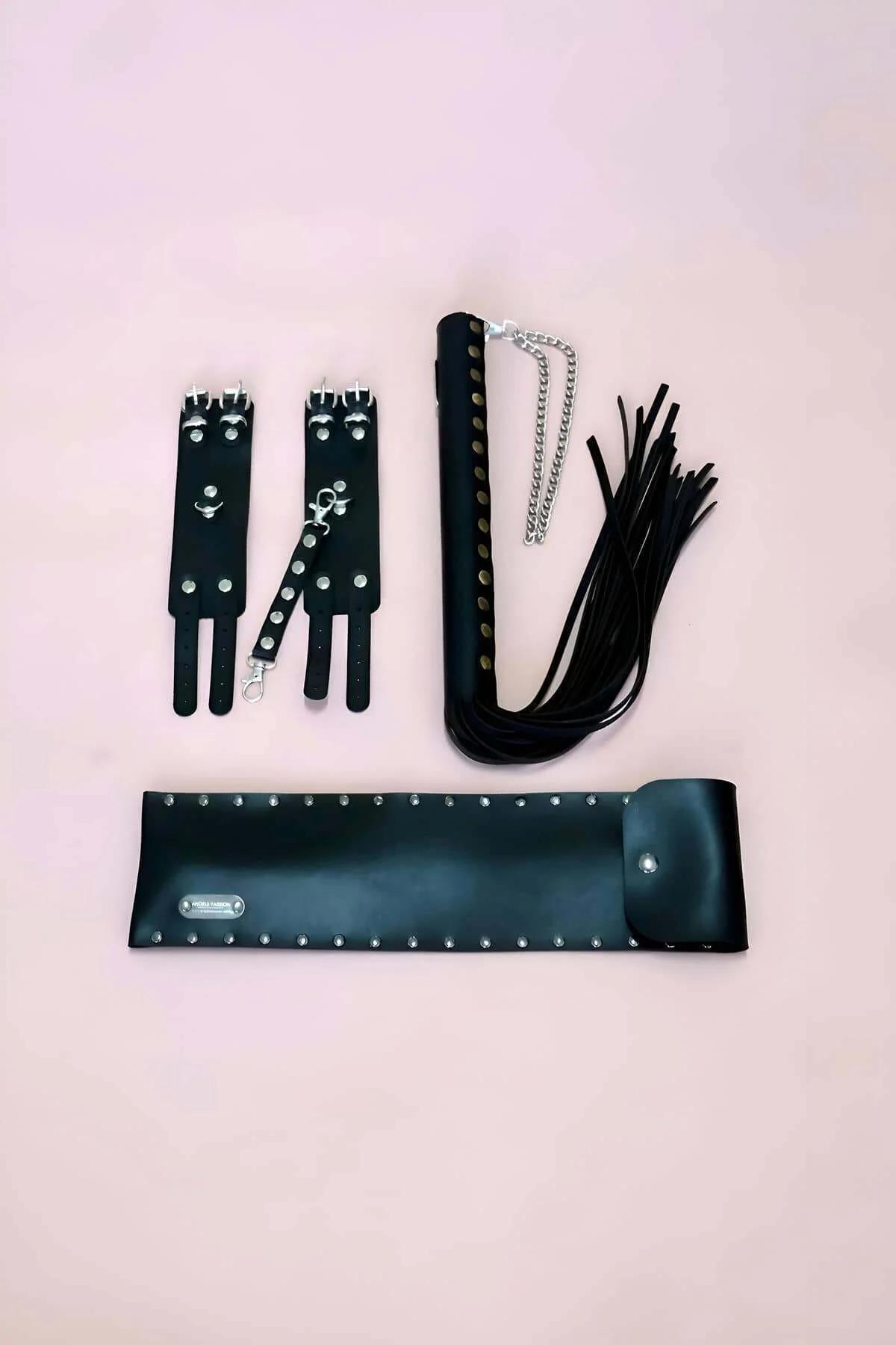 Black vegan leather 3-piece accessory set with studded wrist cuffs, silver chain flogger, and wide studded belt on pink backg