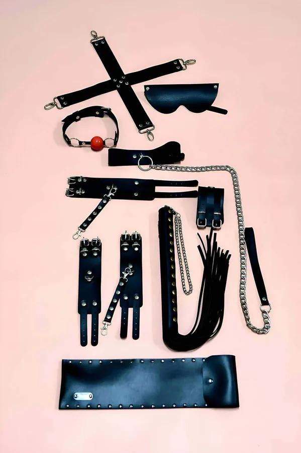 Black vegan leather 7-piece restraint accessory set with silver hardware laid out flat on pink background