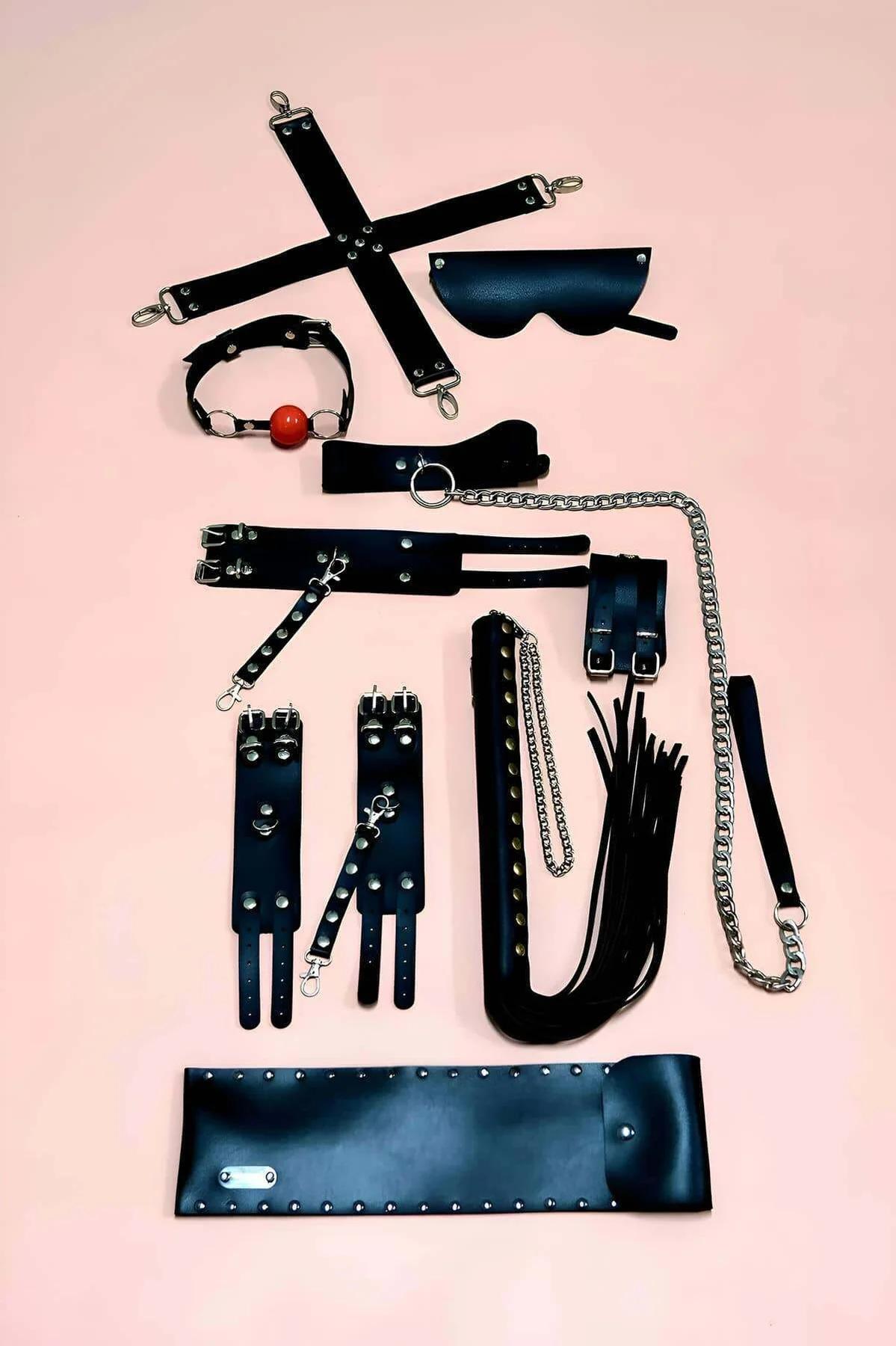 Black vegan leather 7-piece restraint accessory set with silver hardware laid out flat on pink background