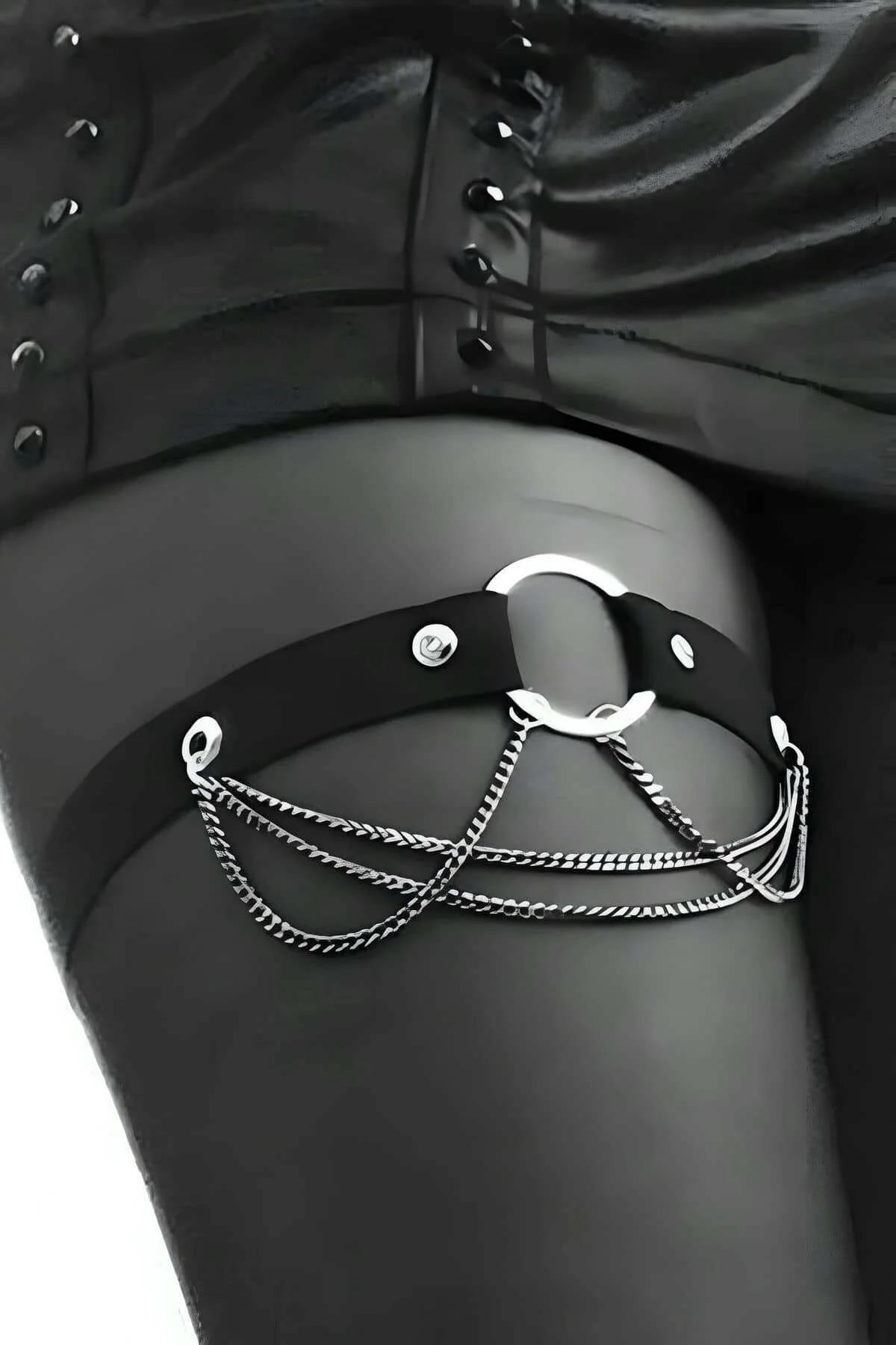 Black faux leather garter harness with silver O-ring and triple chain draping worn on upper thigh over sheer black stockings