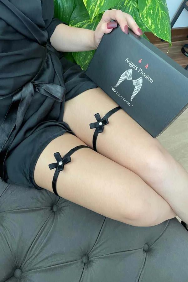 Black satin leg garters with pearl-centered bow detail worn on upper thigh, elastic ribbon band, pair view