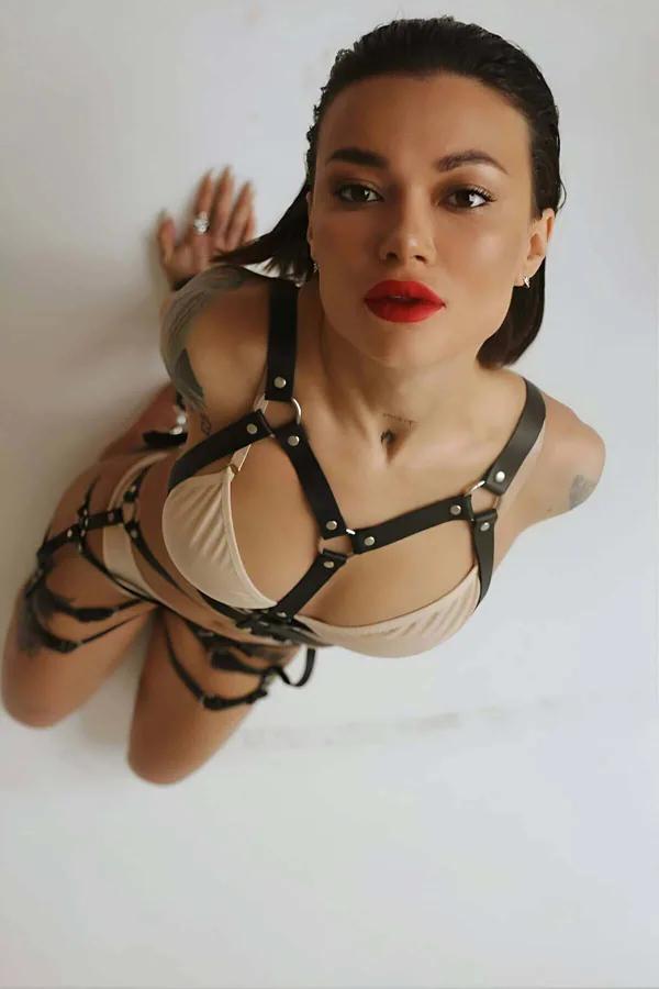 Black faux leather full body harness and thigh strap set with silver O-rings worn over nude bralette, front view