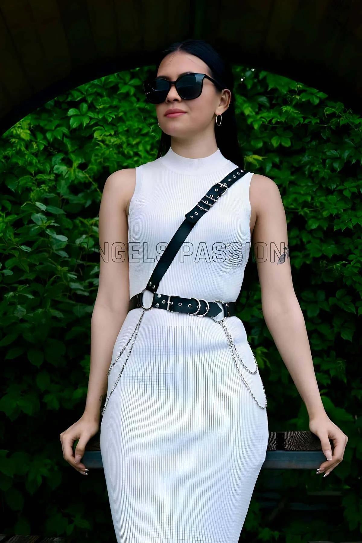 Black faux leather asymmetric chest harness with wide waist belt silver O-rings and draping chains worn over white dress