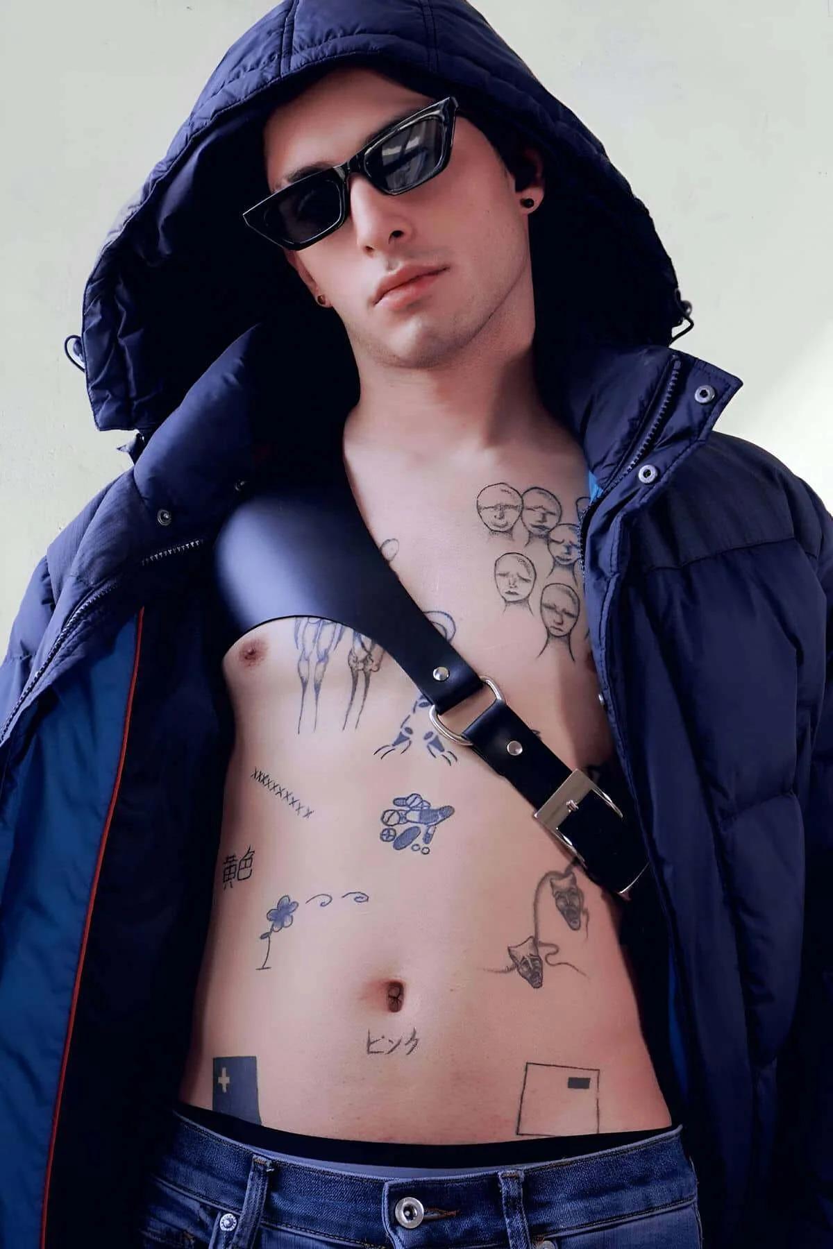 Black faux leather single shoulder harness with silver buckle worn diagonally across bare chest under open navy puffer jacket