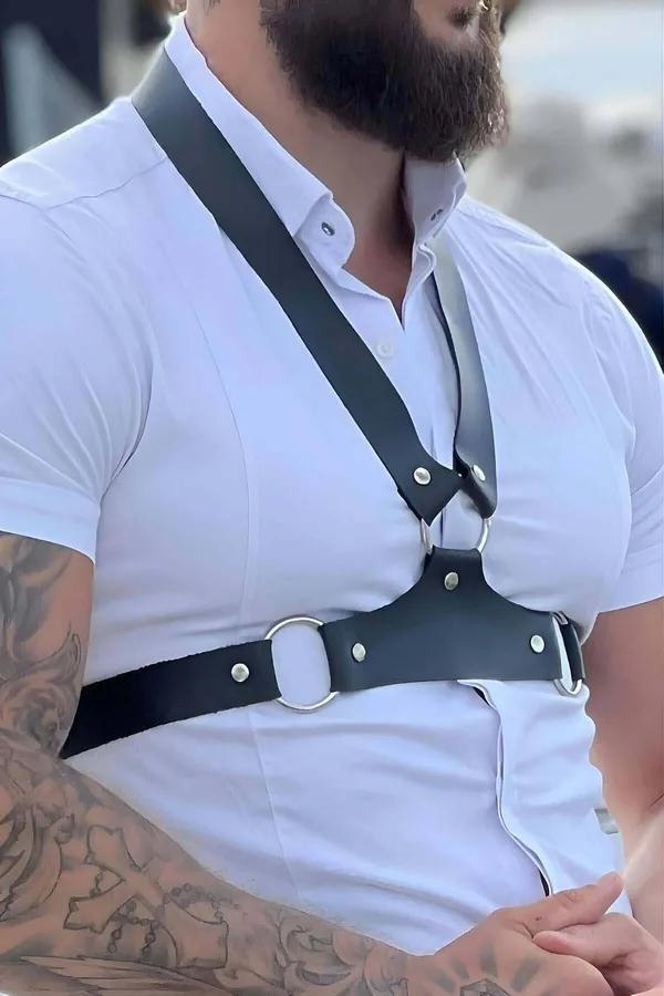 black faux leather chest harness with silver O-ring center worn over white shirt, front view showing cross-body strap and riv
