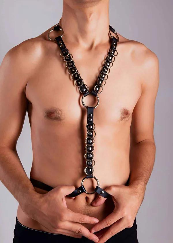 Black vegan leather chest harness with continuous silver O-ring chain running from collar to waist, worn by male model
