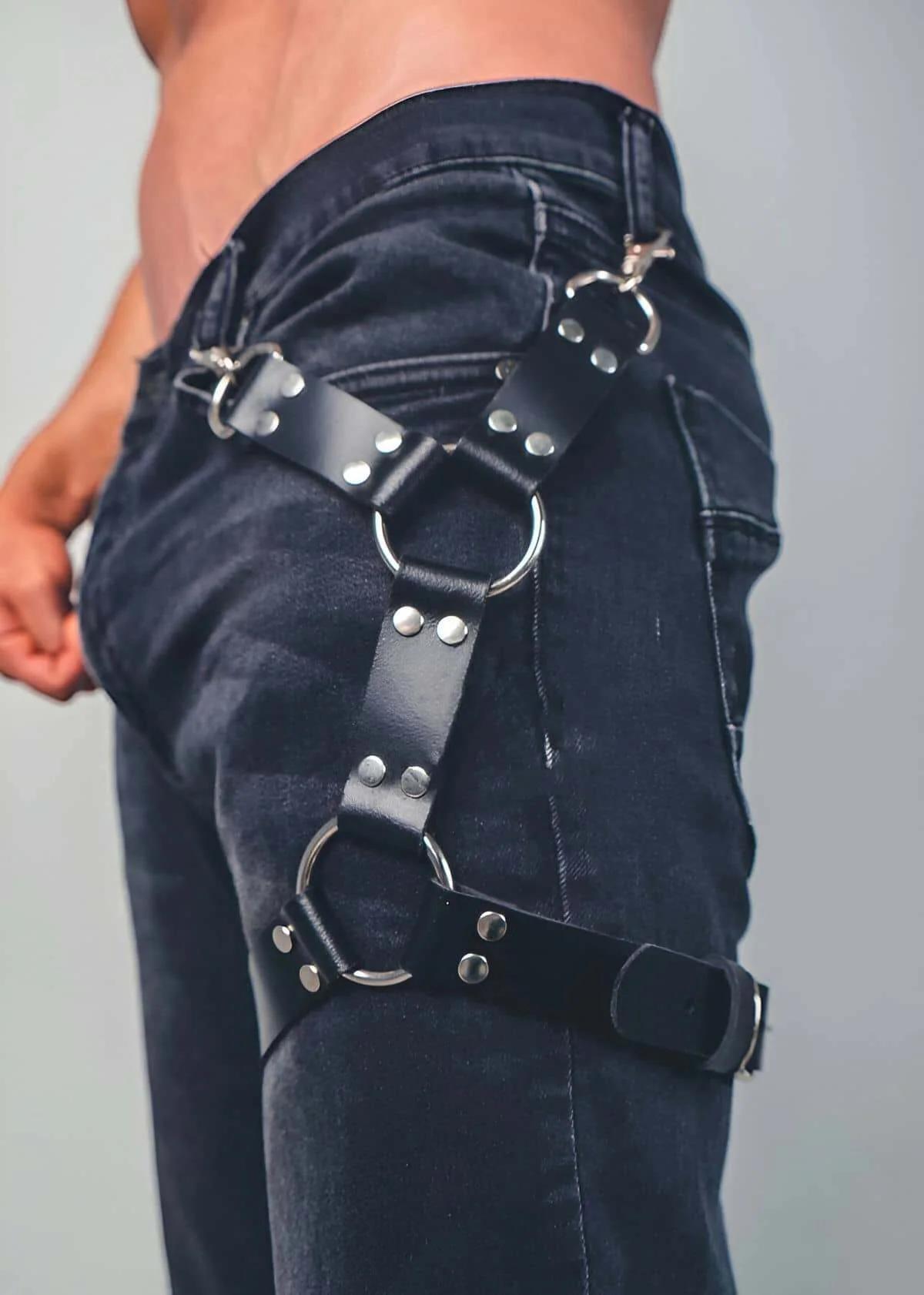 Black faux leather thigh harness with two silver O-rings and rivet-studded straps worn over dark denim jeans